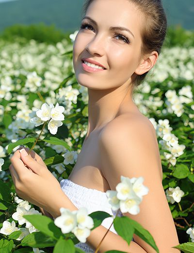 Smiling woman in white dress holding jasmine flower in blooming jasmine field under sunny sky, symbolizing the soothing and antioxidant-rich Jasmine extract in Oryskincare's rejuvenating skincare products for hydrated, youthful skin.