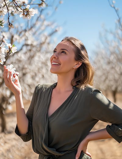 Woman in a blooming almond orchard with radiant, nourished skin, symbolizing the gentle moisturizing and soothing benefits of sweet almond oil in skincare.
