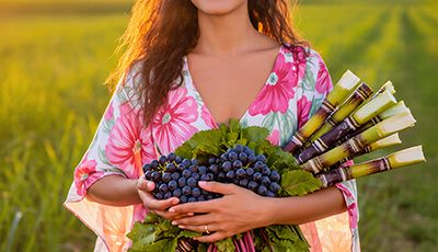 Smiling woman in floral dress holding fresh sugarcane, grapes, and beets in sunny field, symbolizing natural sources of Glycolic Acid for Oryskincare exfoliating and renewing skincare products.